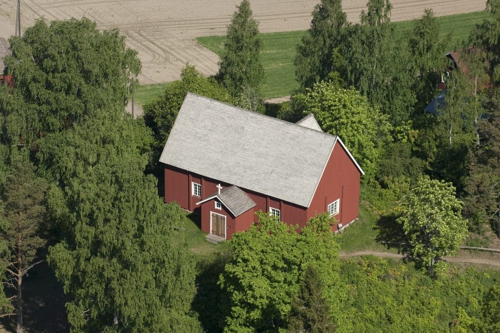 Lopen punamullan värinen vanha puinen kirkko pelto- ja puistomaisemassa.